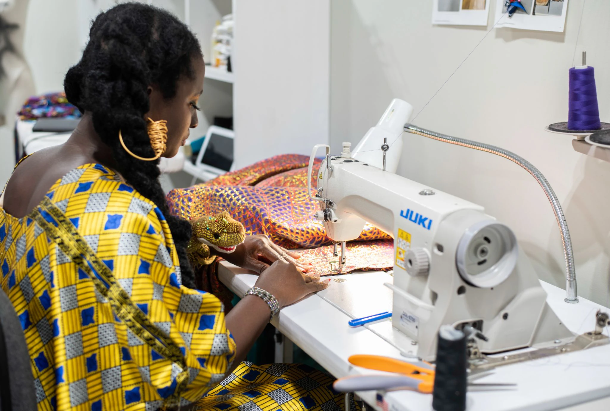 A woman with long black braids, gold hoop earrings, and a blue and yellow patterned dress sits at a sewing table making a garment.