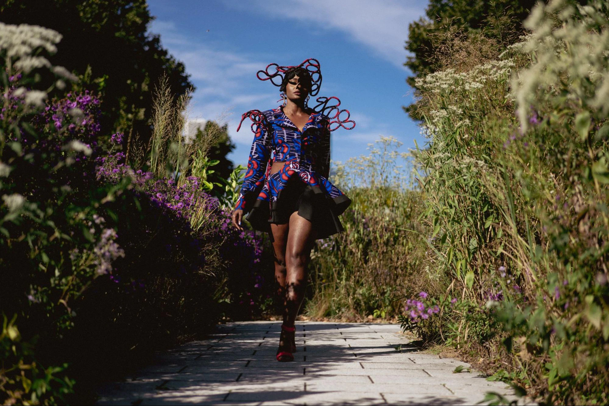 A model walks on a path through a garden wearing a vibrantly colored garment and headpiece.