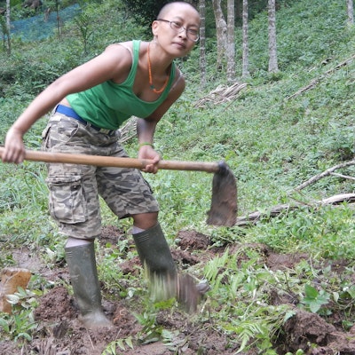 A women in a green tank top and boots, holding a garden hoe