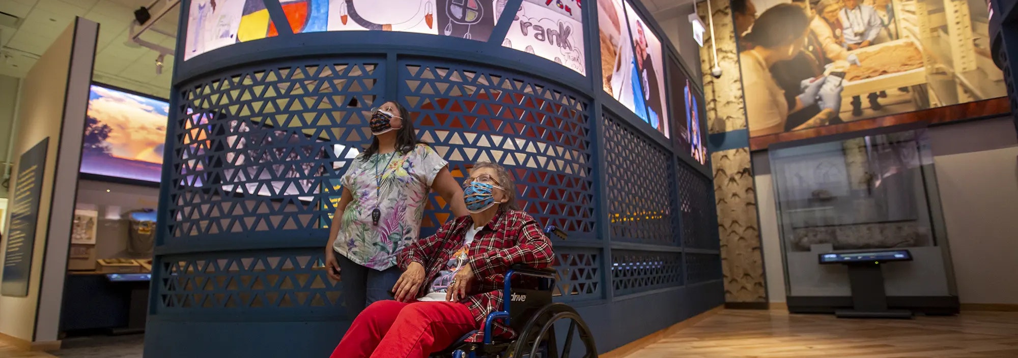 Visitors inside the Native Truths: Our Voices, Our Stories exhibition look up at an exhibit. Both visitors are wearing medical face masks. One sits in a wheelchair.