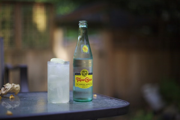 A Ranch Water cocktail in a highball glass and a bottle of Topo Chico on a tabletop, outdoors