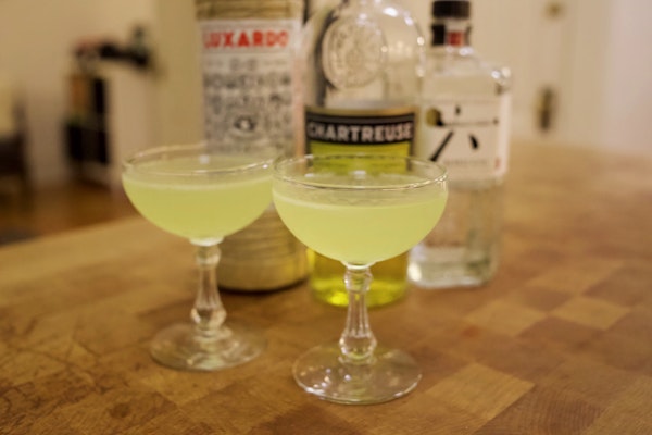 Two green cocktails in coupe glasses in front of bottles of liquor 