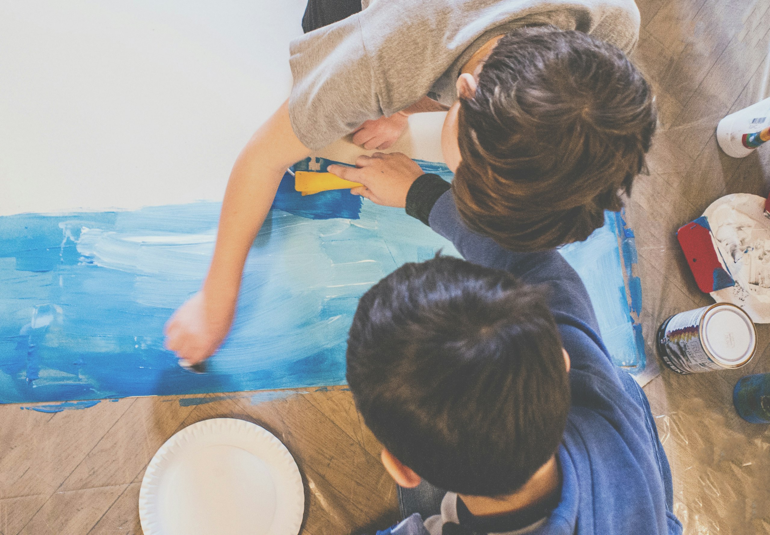 Dvě děti malují na papír na zemi modrou plochu v jednom z výtvarných kurzů ve Ville Pellé.