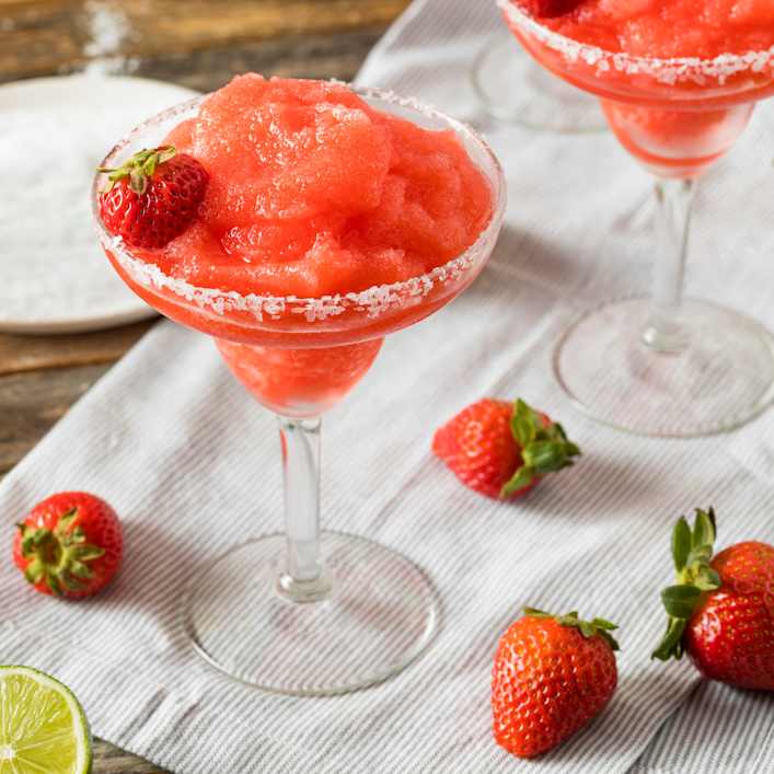 Cocktailglas mit Erdbeer-Daiquiri steht auf einem Tuch auf einem Tisch, daneben sind Erdbeeren platziert.