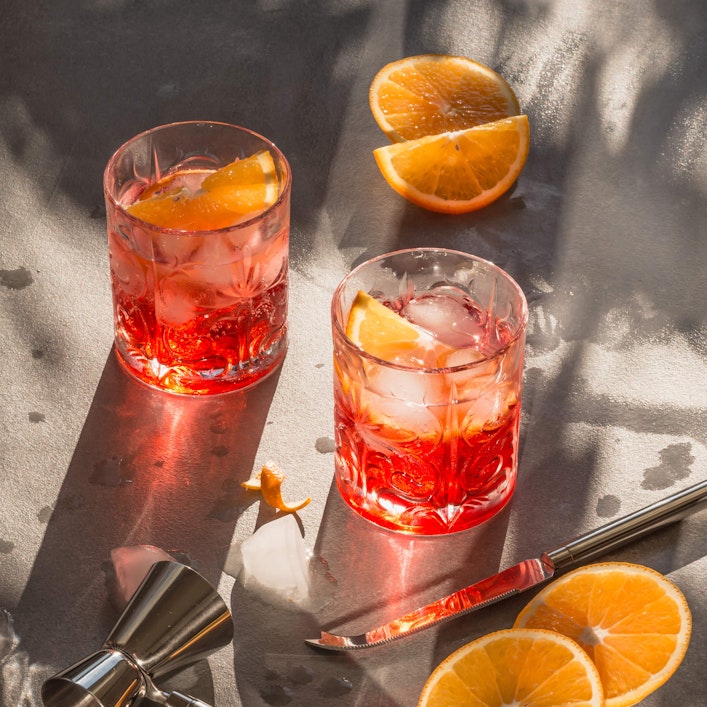 Zwei geschliffene Gläser mit Campari sind mittig platziert. Die schein von schräg oben auf sie herab, wodurch die Gläser Schatten werfen. Um die Gläser herum sind Cocktail-Utensilien und Orangenscheiben platziert.