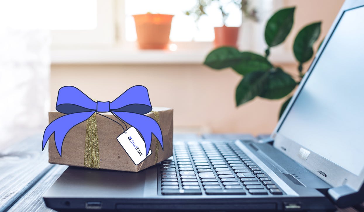 A photo of a blurred laptop screen. On top of the opened laptop screen is a gift box. On top of the gift box is a StartMail brand style illustrated blue ribbon, and a tag with the StartMail logo on it. The image represents privacy gifts for someone who values online security and privacy.