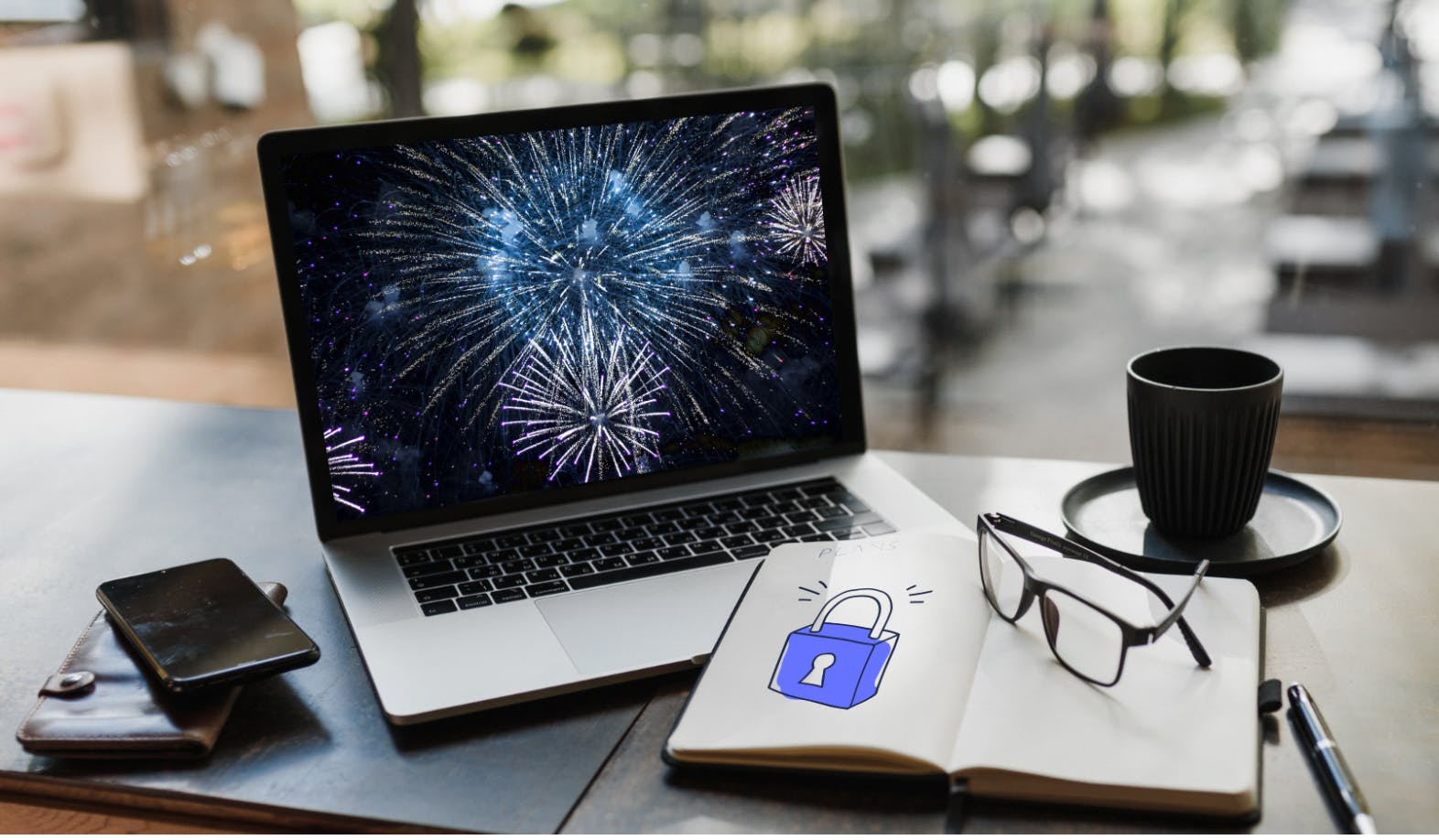 A photograph of a laptop displaying purple and blue fireworks. On the left side of the laptop is a wallet and mobile laying on a table. On the right side of the laptop is a cup. In the front of the laptop is a notebook, with an illustration of a lock in the StartMail brand style. On top of the notebook there are reading glasses.