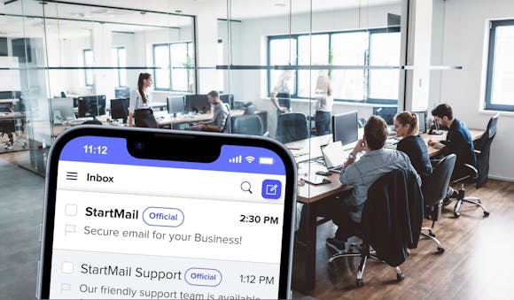 An office setting with employees working at desks and computers. In the foreground, a smartphone screen shows the StartMail inbox with messages from 'StartMail', emphasizing secure email for business and support availability.
