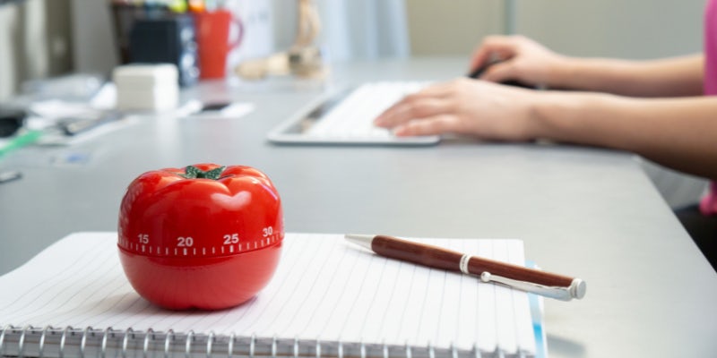 Close-up of a red tomato-shaped Pomodoro timer on a notebook, with a pen and a person typing on a keyboard in the background.
