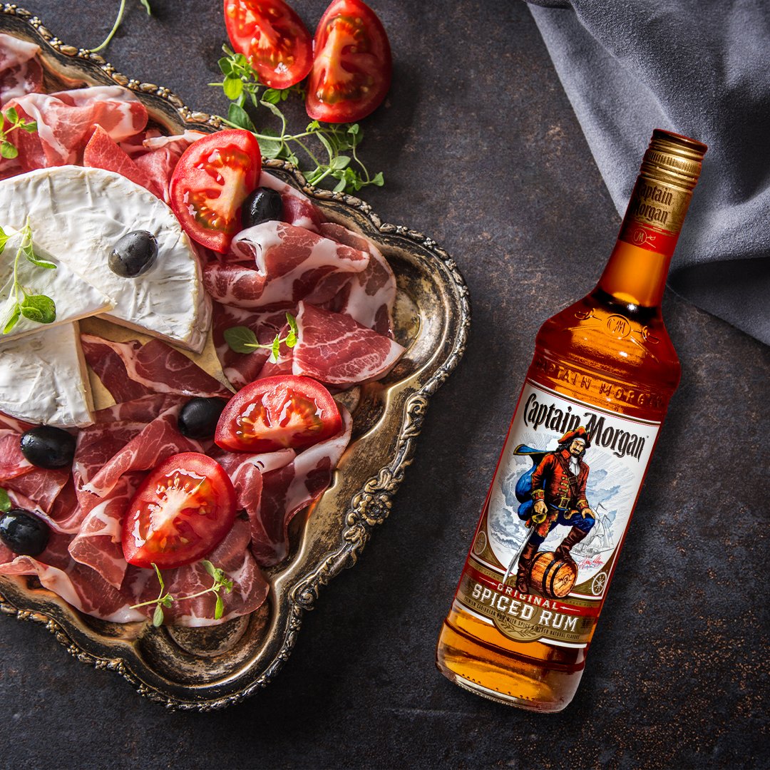 A bottle of Captain Morgan spiced rum lays across a table beside a plate of cold cuts