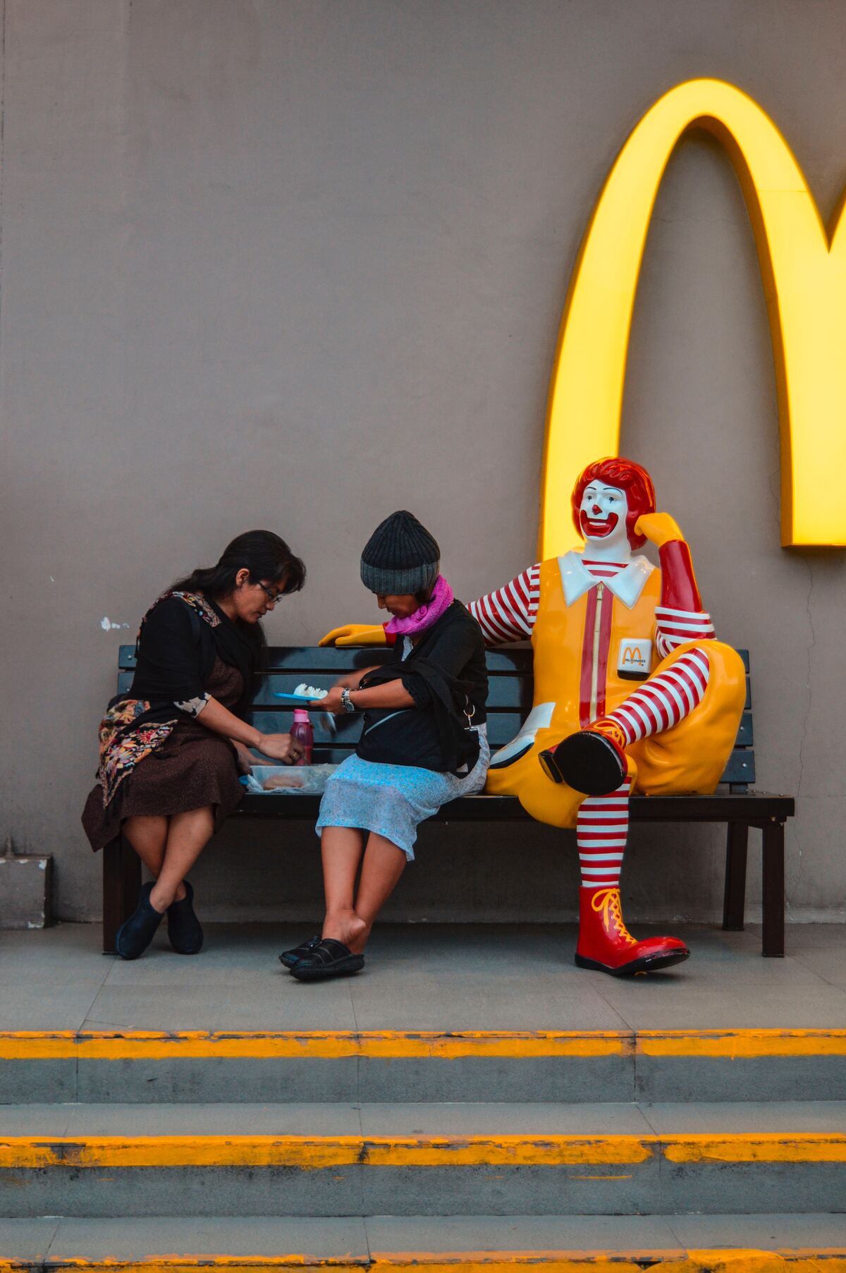 Two ladies share a meal on a bench with Ronald Mcdonald beside them.
