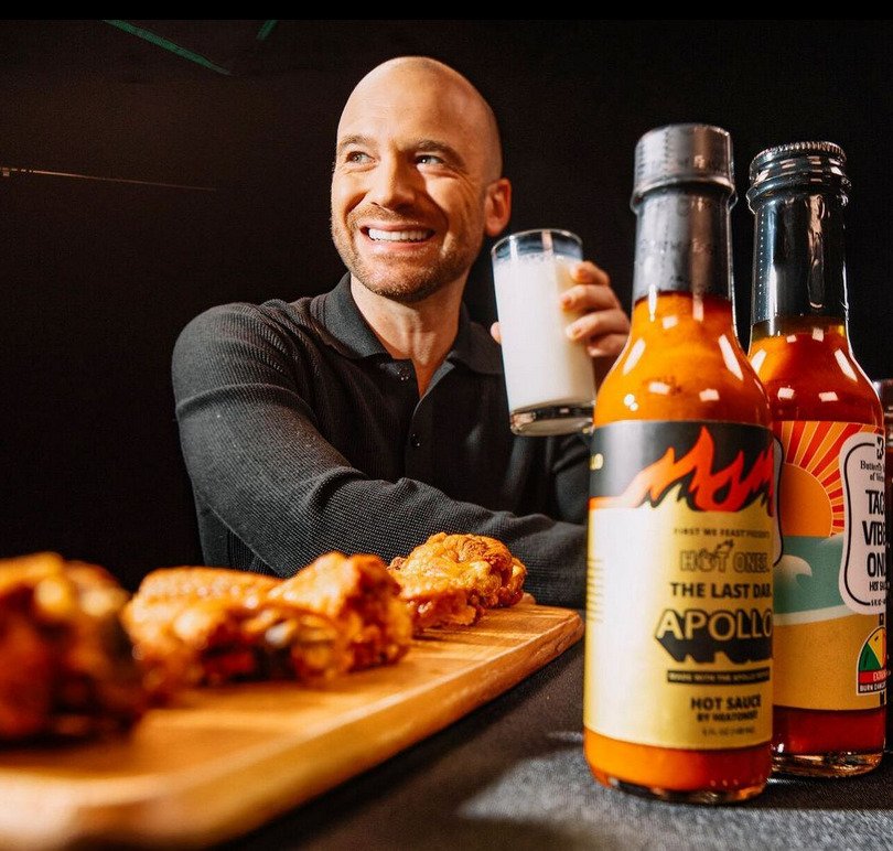 A bald man holding a glass of milk looks to the side as he smiles. In front of him are chicken wings and two bottles of hot sauce