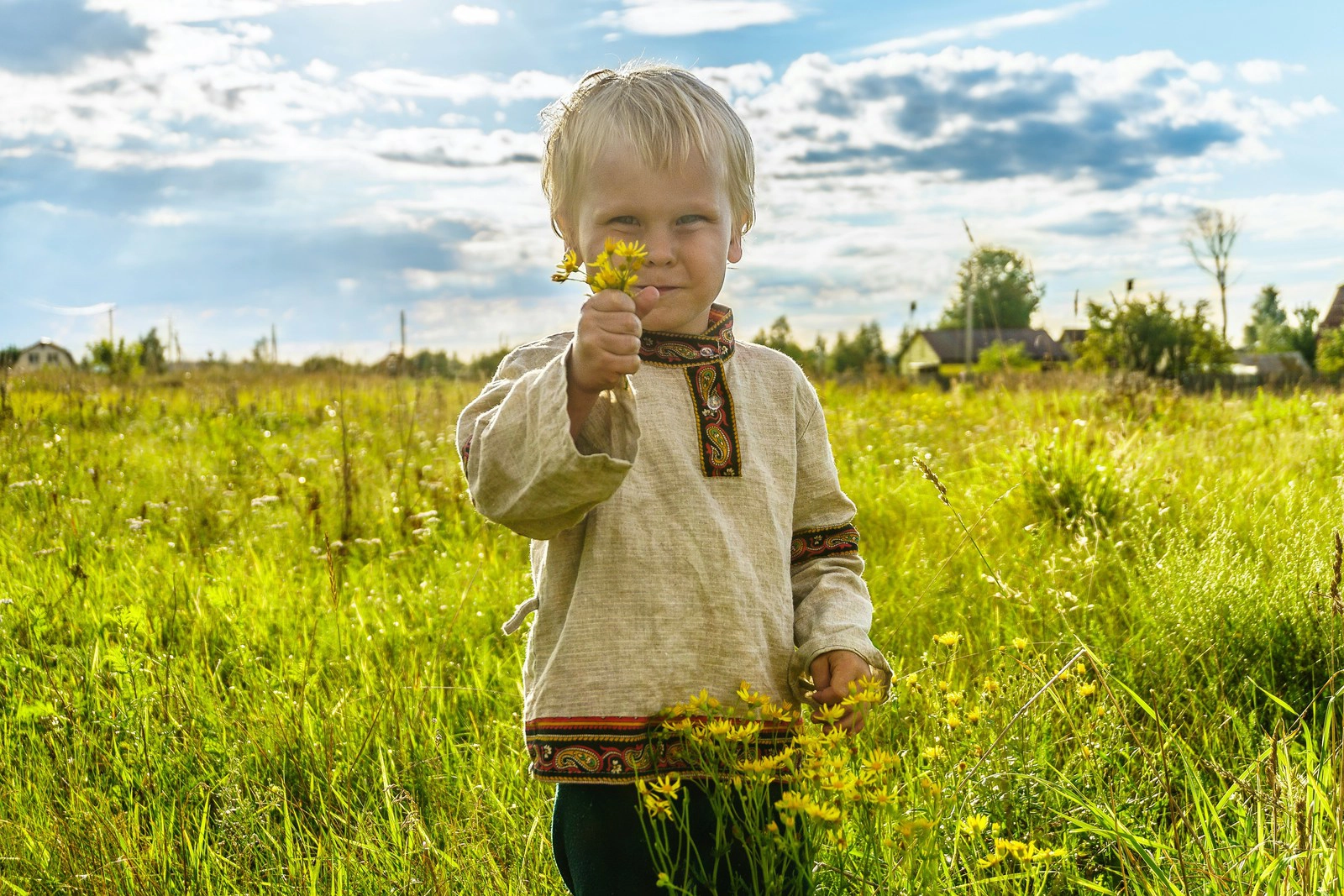 A boy in a gray sweater standing on a green grass field during the daytime smiles as he holds a yellow flower