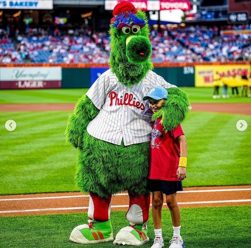 A boy wearing a red shirt stands happily beside a green furry mascot