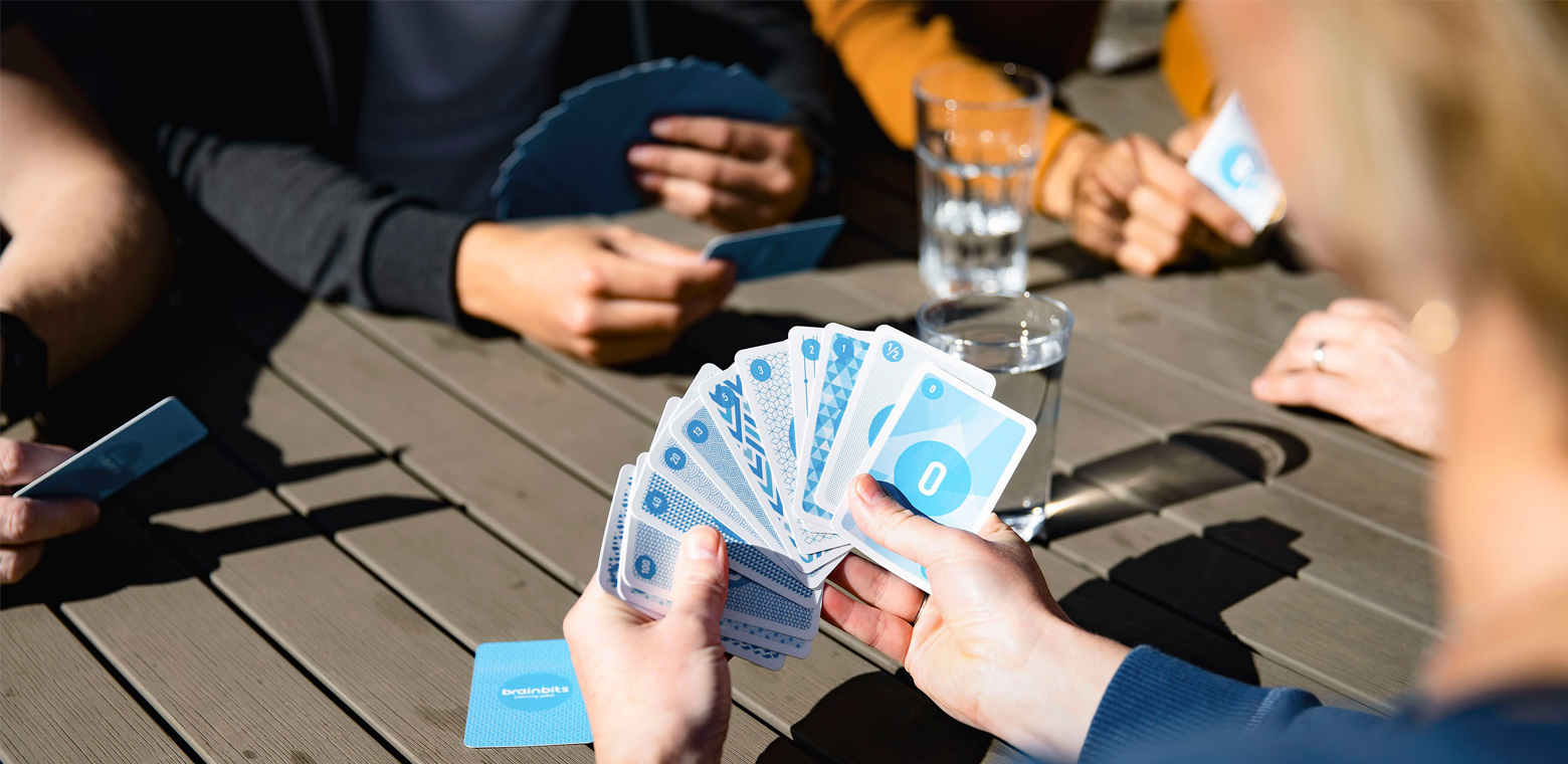 Hand hält Planning-Poker-Karten mit verschiedenen Zahlen darauf, im HG weitere Handpaare mit Karten, die auf einem Tisch aufgestützt sind