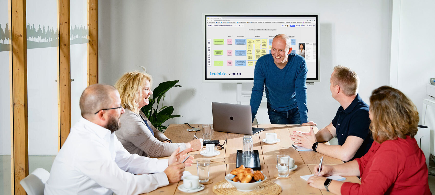 Vier Personen sitzen an einem Tisch. Eine steht vor ihnen und erklärt lächelnd etwas. Auf dem Bildschirm im Hintergrund ein Ausschnitt aus dem Tool Miro.