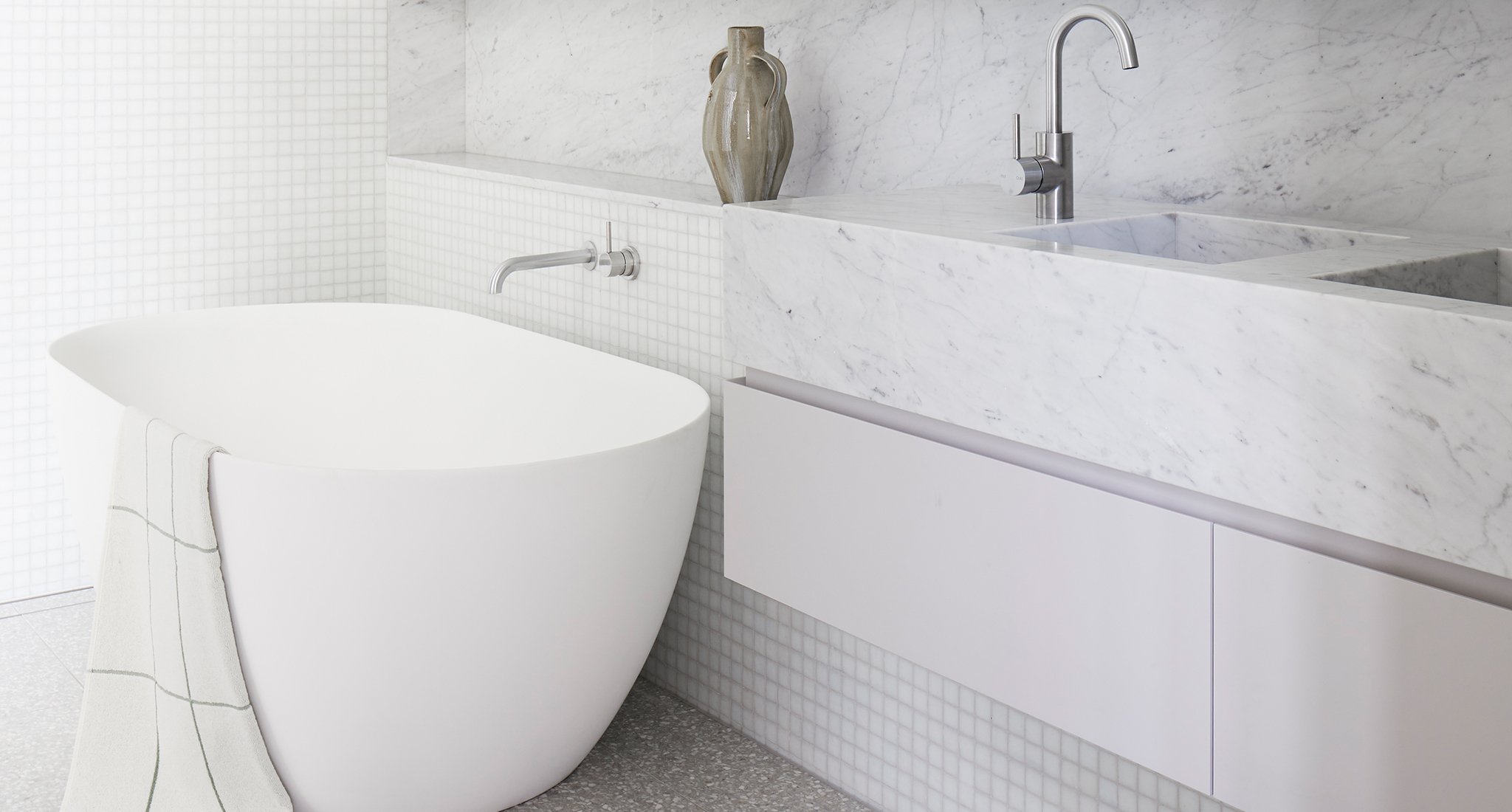 High end photography shot by Dave Wheeler of luxury white bathroom featuring grey terrazzo floor tiles and small white mosaic tiles throughout. Custom vanity with marble bench top which integrates into the wall. The bathrooms has brushed nickel tap ware throughout and a large white soaking tub. 