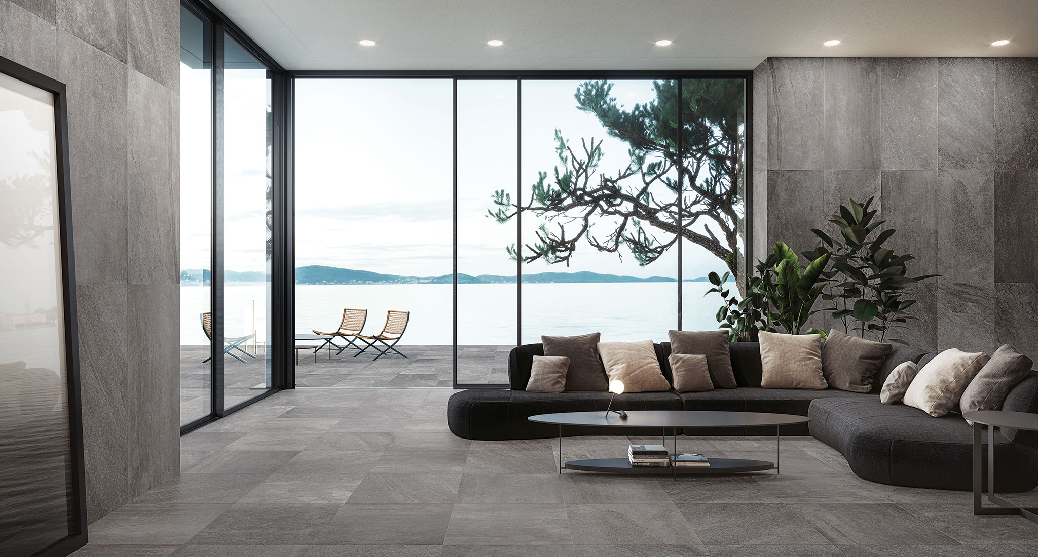 Deep Grey large format tiles featured on the floor and walls of the room. Large glass sliding doors lead onto a waterfront deck with deck chairs and a view of a lake. Inside, we see a large charcoal coach with cushions and a coffee table. On the left, a glimpse of a mirror reflect the lake and trees in the distance.