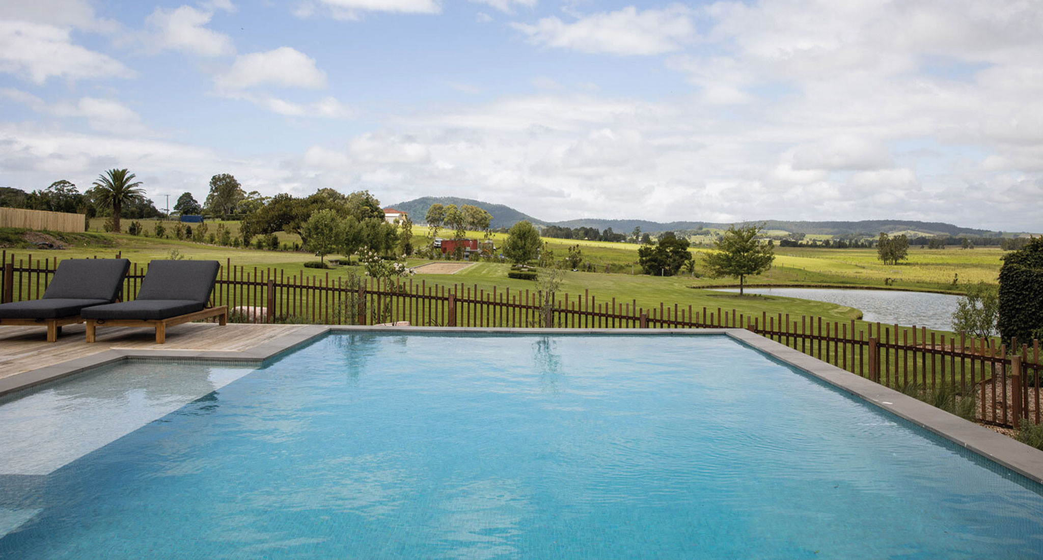 Landscape shot of pool facing out into the Australian countryside. Hills and green grass in the distance and a lake. The pool has crystal clear water and two deck chairs are on the left.