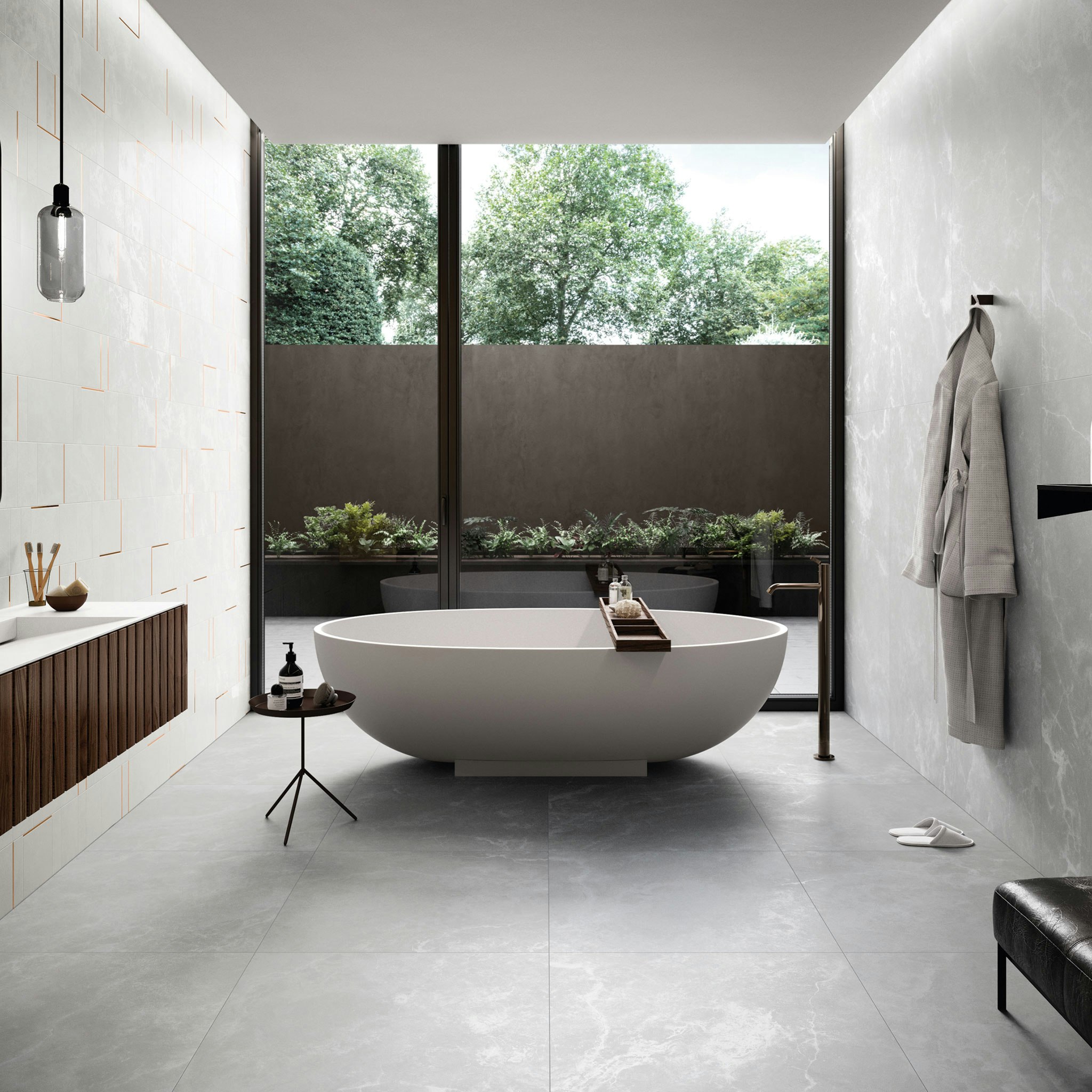 Sparkling light grey tiles on walls and floor in a large format. A bath sits in the centre of the room which looks out into a modern courtyard. On the left, we see a feature mosaic in the same tile with a bronze insert. A glimpse of a vanity and pendant can also be seen on the left. On the right a robe is hanging off the wall and we see a glimpse of a bench. The daylight shines off the floor tiles.