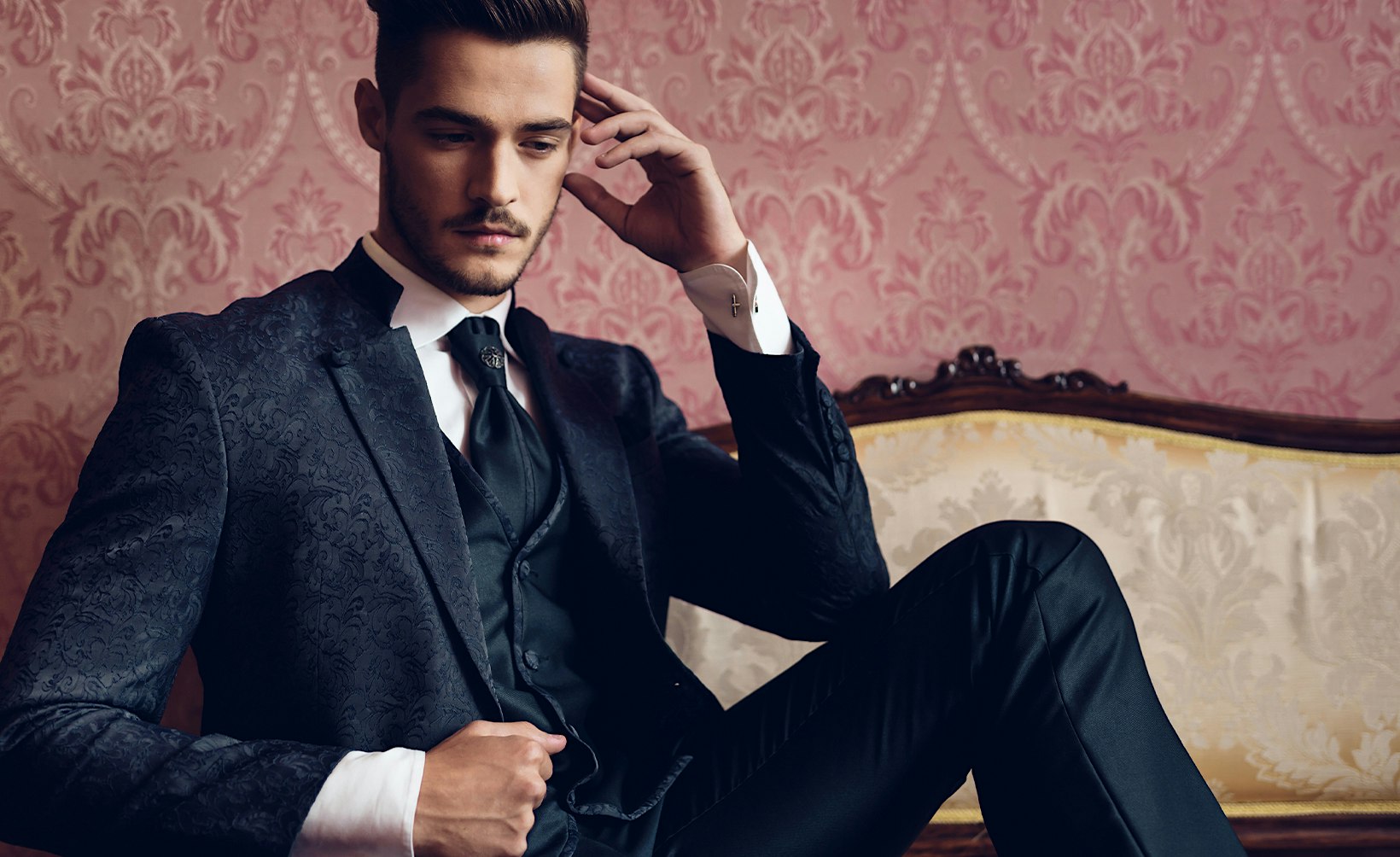 man in a black suit sitting on a couch with his hand on his head