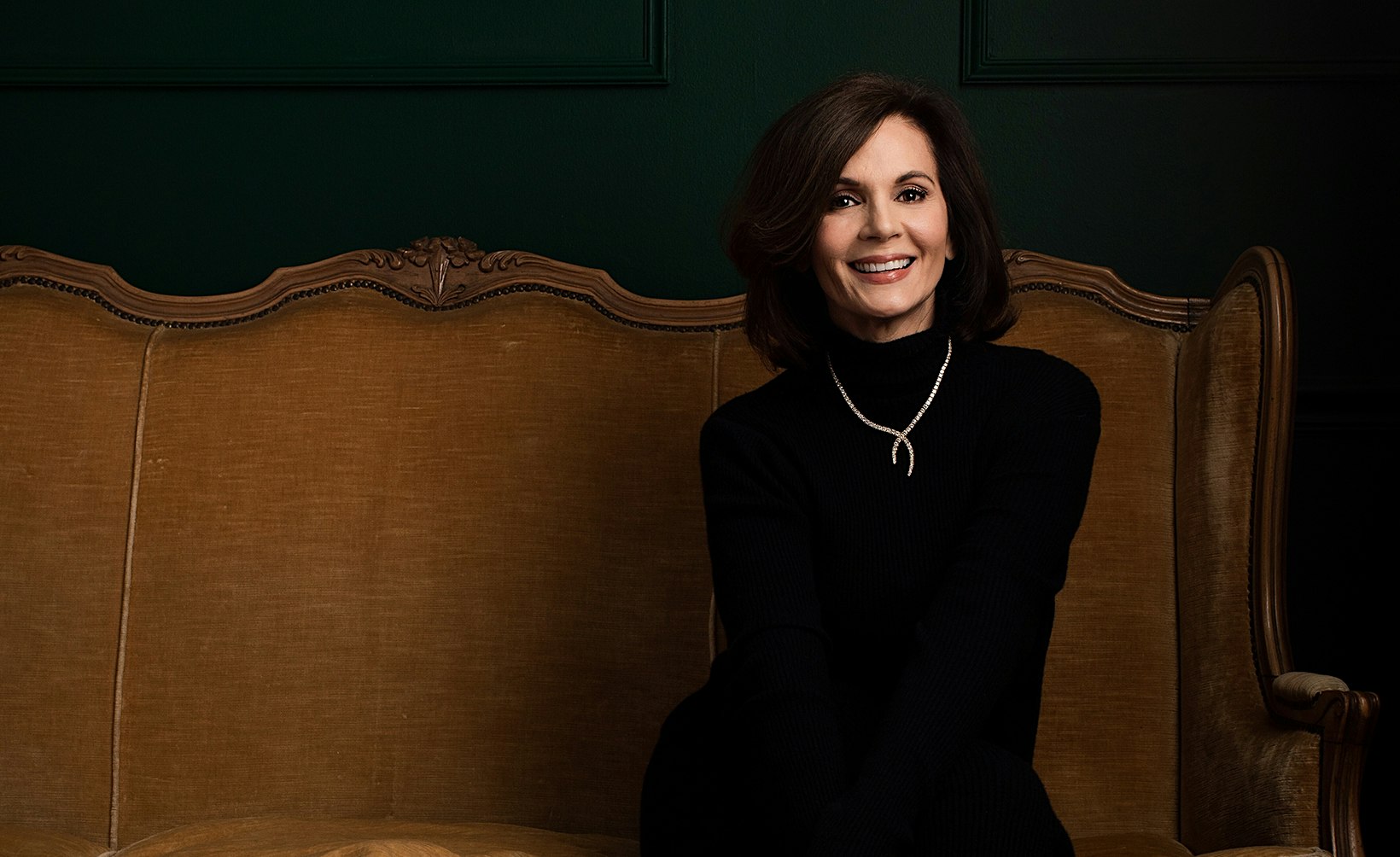 a woman sitting on a couch with a necklace and black turtleneck on