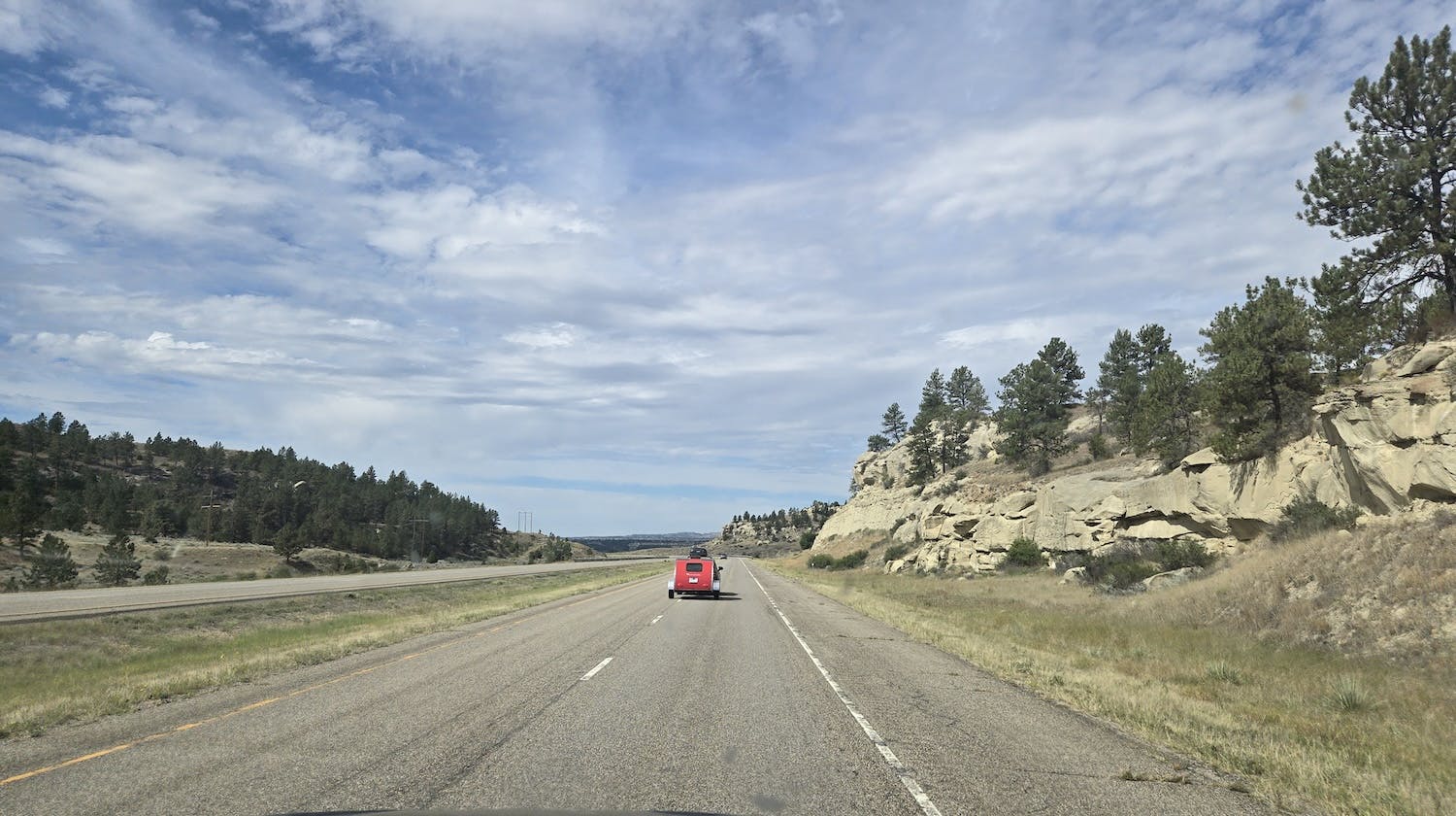 montana highway with rocky hills left and right and a car towing a small red camper ahead