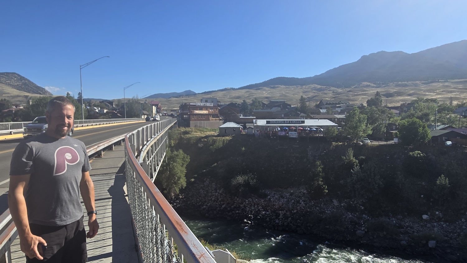Jason posing on walking bridge over the Yellowstone River with restaurants in the background atop a steep cliff