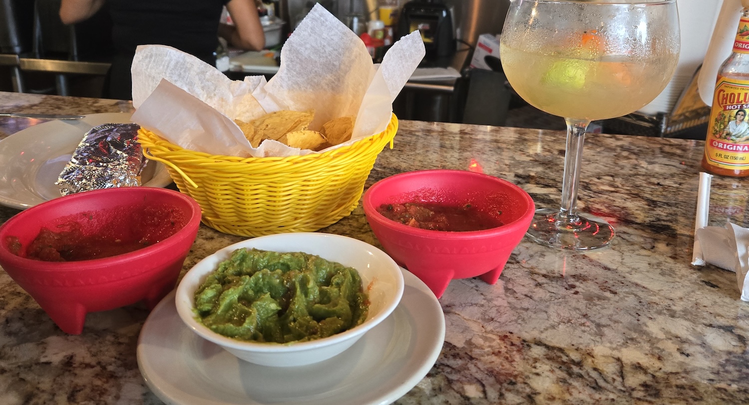 a bowl of guacamole, two red dishes of salsa on each side, bowl of chips and margarita