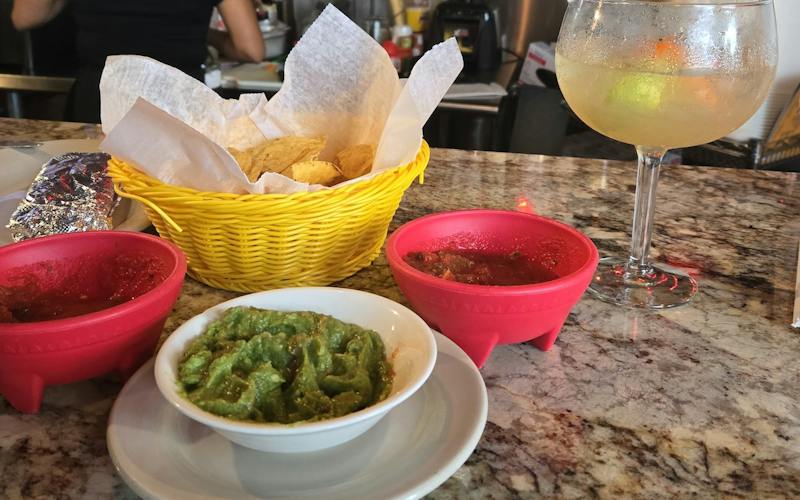 a bowl of guacamole, two red dishes of salsa on each side, bowl of chips and margarita