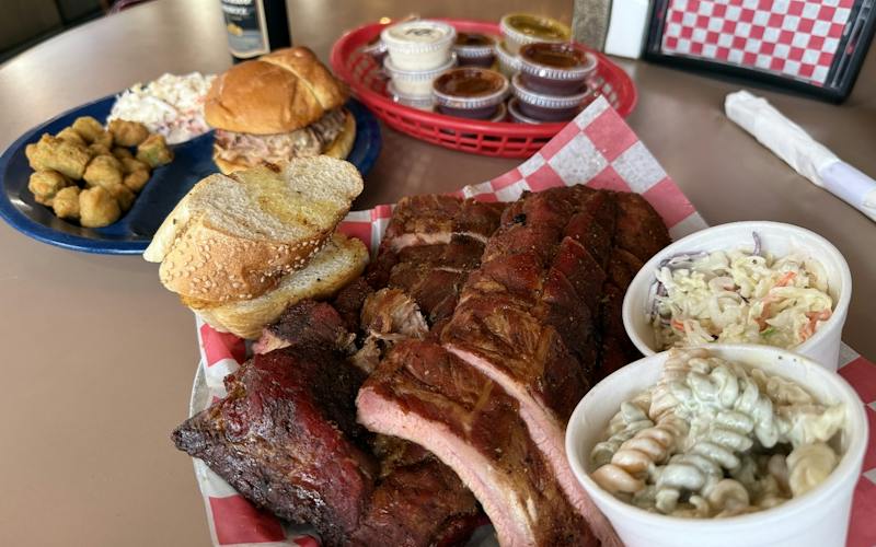 a plate of BBQ ribs with sides of pasta salad and coleslaw, another sandwich sides and sauces in background
