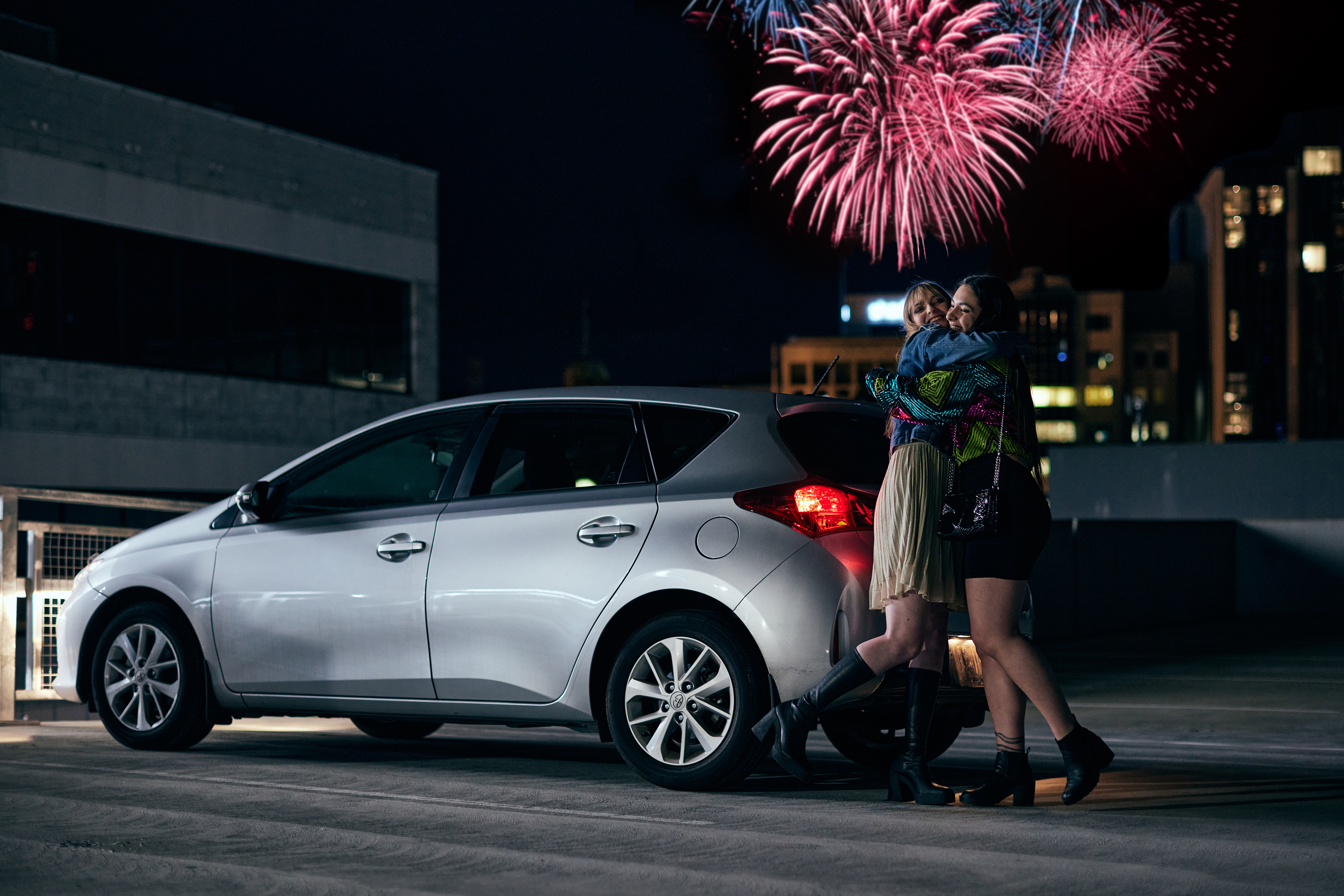 Girls hugging in car park with fireworks in the background.