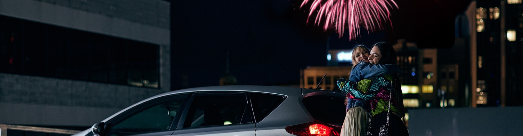 Girls hugging in car park with fireworks in the background.