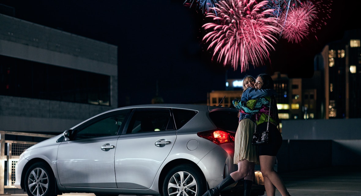 Girls hugging in car park with fireworks in the background.