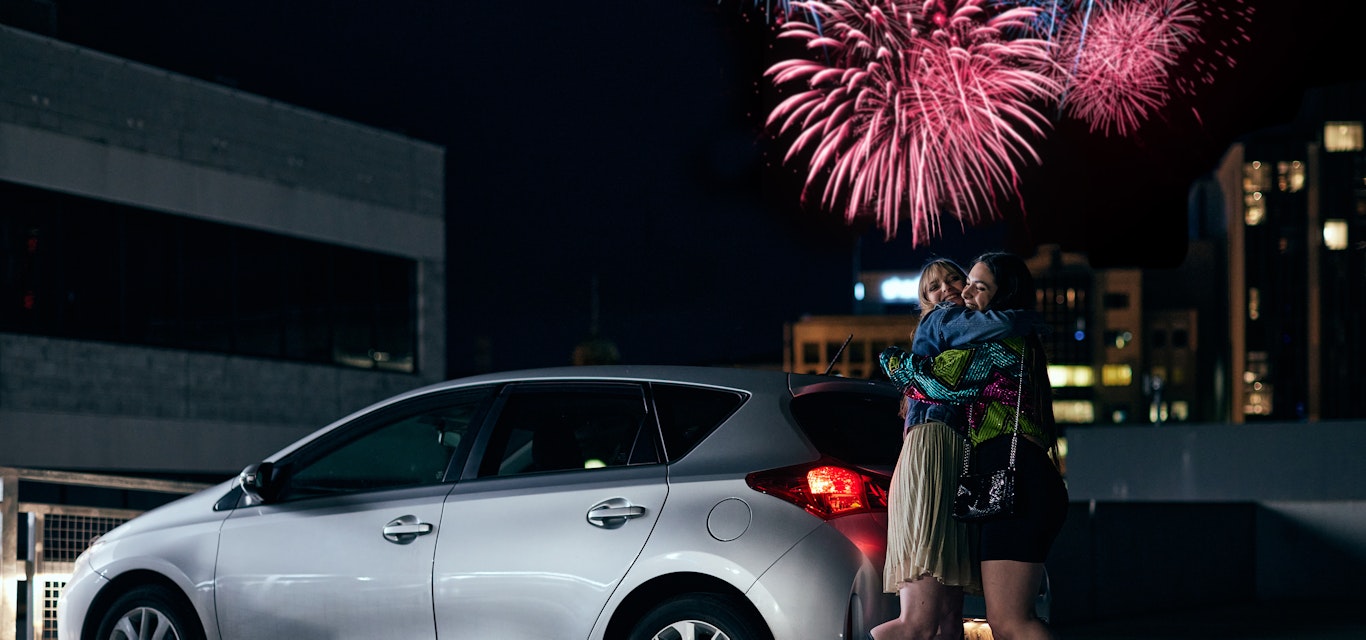 Girls hugging in car park with fireworks in the background.