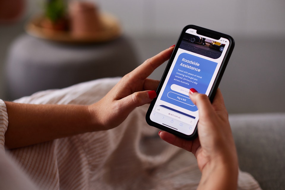 A woman's hands hold a mobile phone with a screen on the RACT Roadside page.