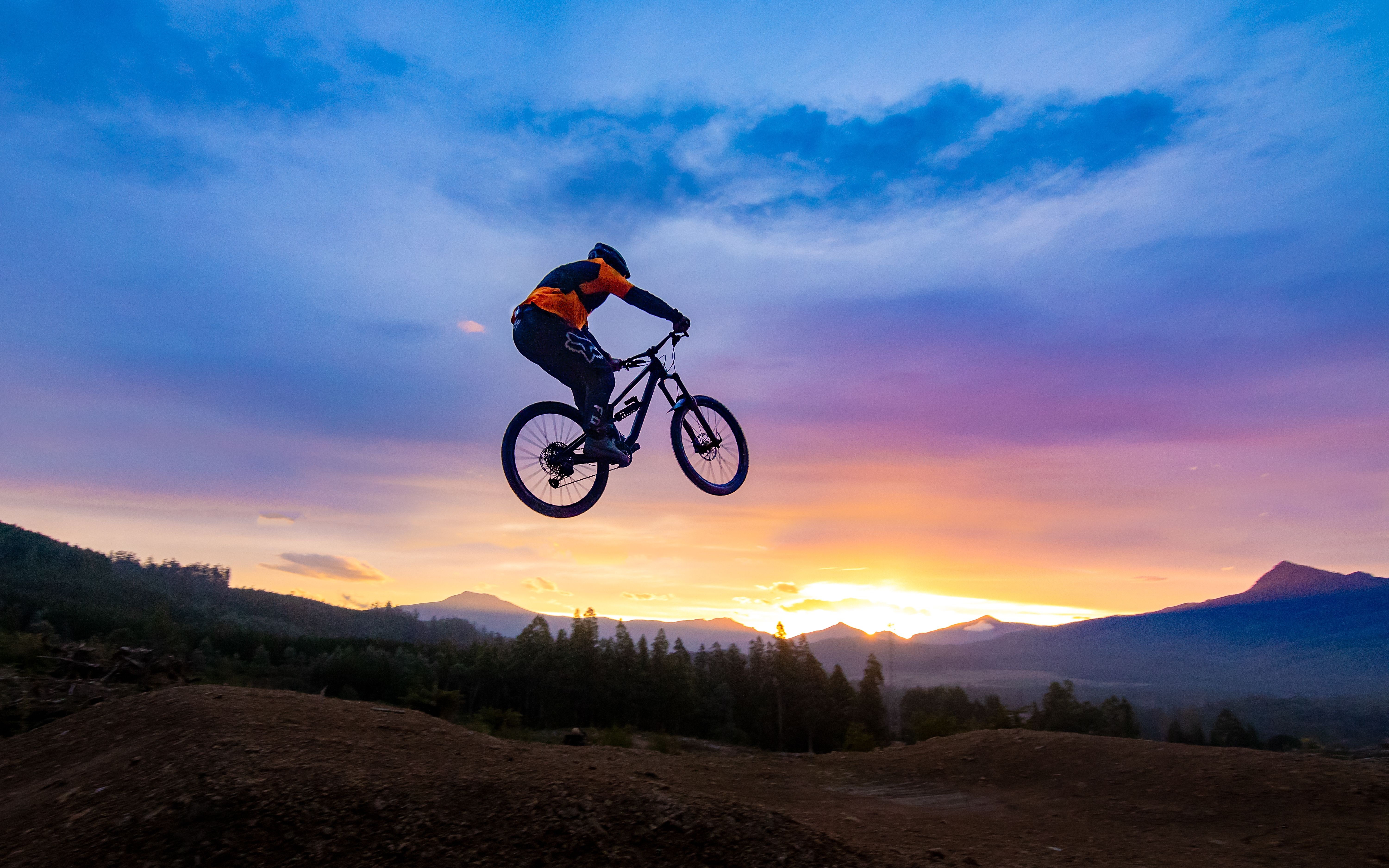A bike rider flies through the air with a beautiful sunset over mountains behind them
