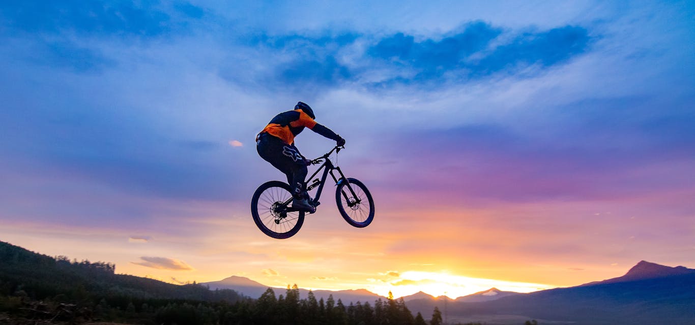 A rider chooses adventure at Maydena Bike Park A bike rider flies through the air with a beautiful sunset over mountains behind them