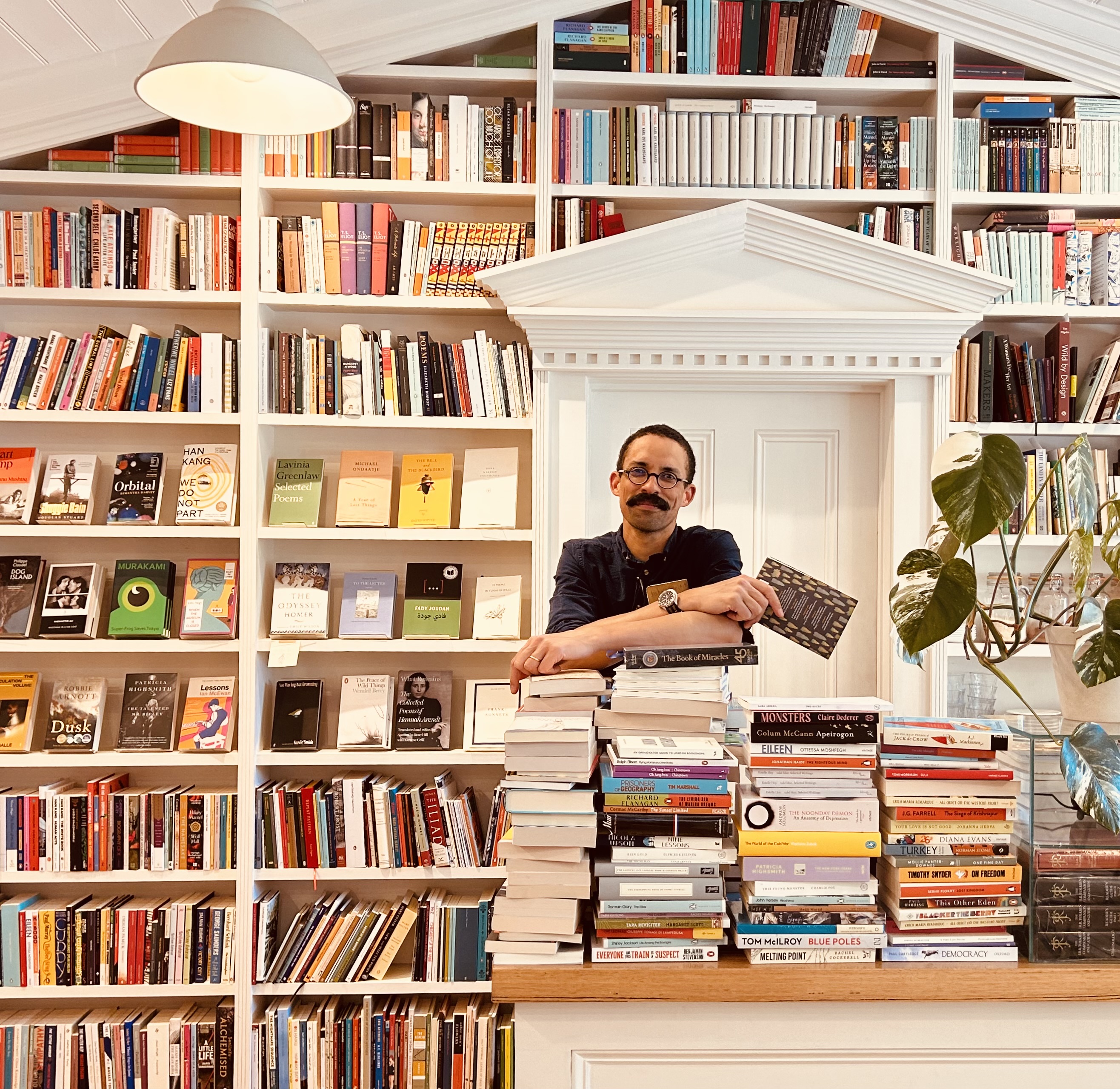 A man with a moustache leans on a pile of books within a small bosy bookshop