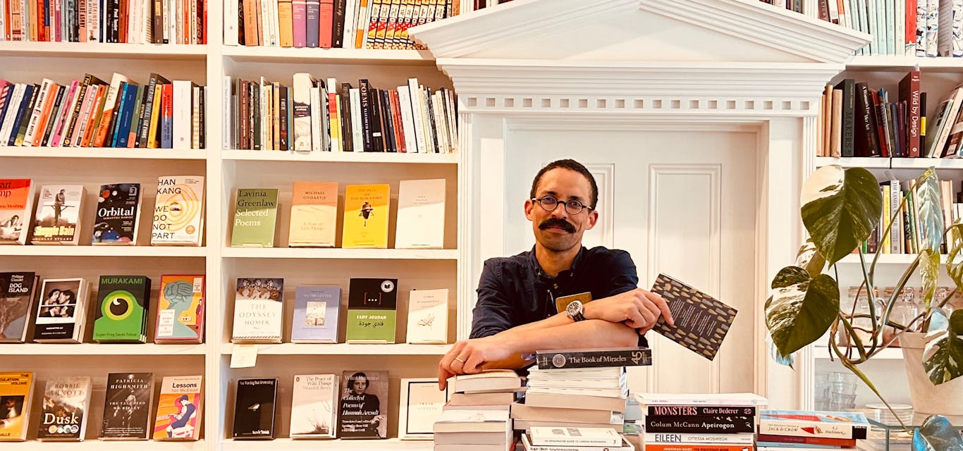 Alexander Okenyo in his cosy Black Swan Bookshop in New Norfolk A man with a moustache leans on a pile of books within a small bosy bookshop