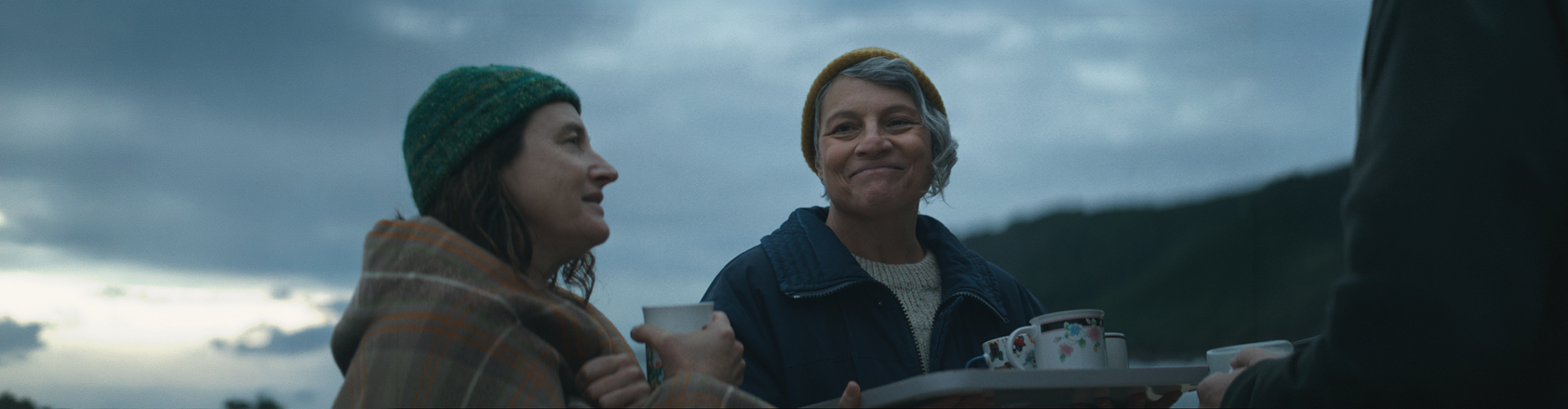 Two women stand together sharing a smile over a tray of tea cups being handed out among their community