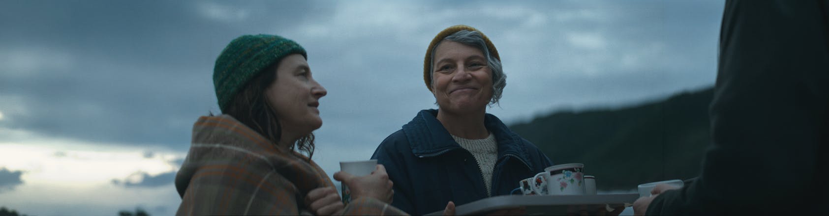 For each other Two women stand together sharing a smile over a tray of tea cups being handed out among their community