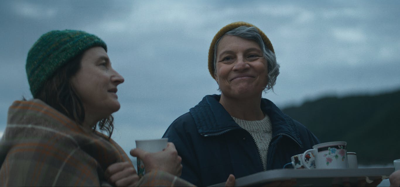 For each other Two women stand together sharing a smile over a tray of tea cups being handed out among their community