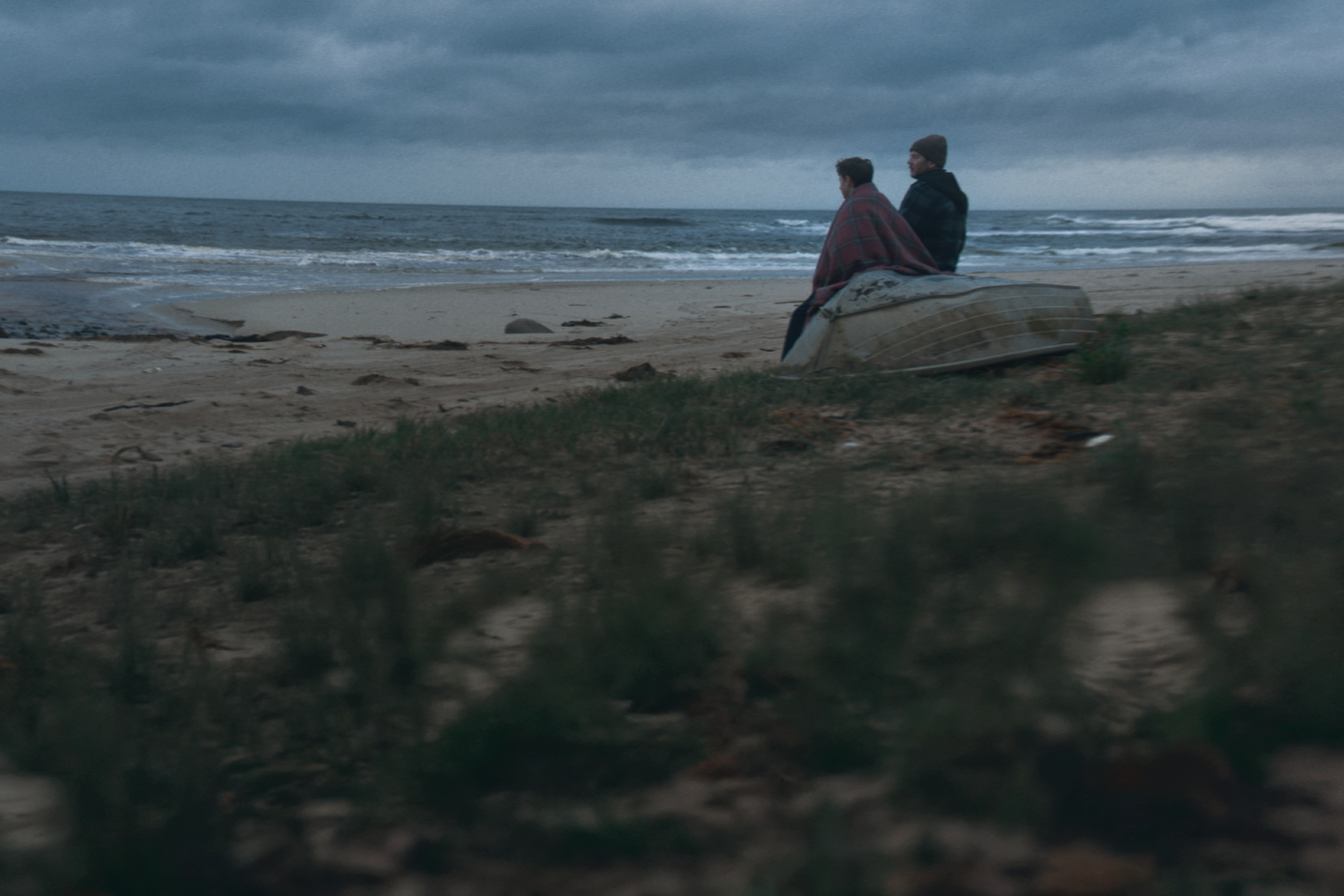 A man and a boy sit on an upturned dinghy on a wild, overcast beach