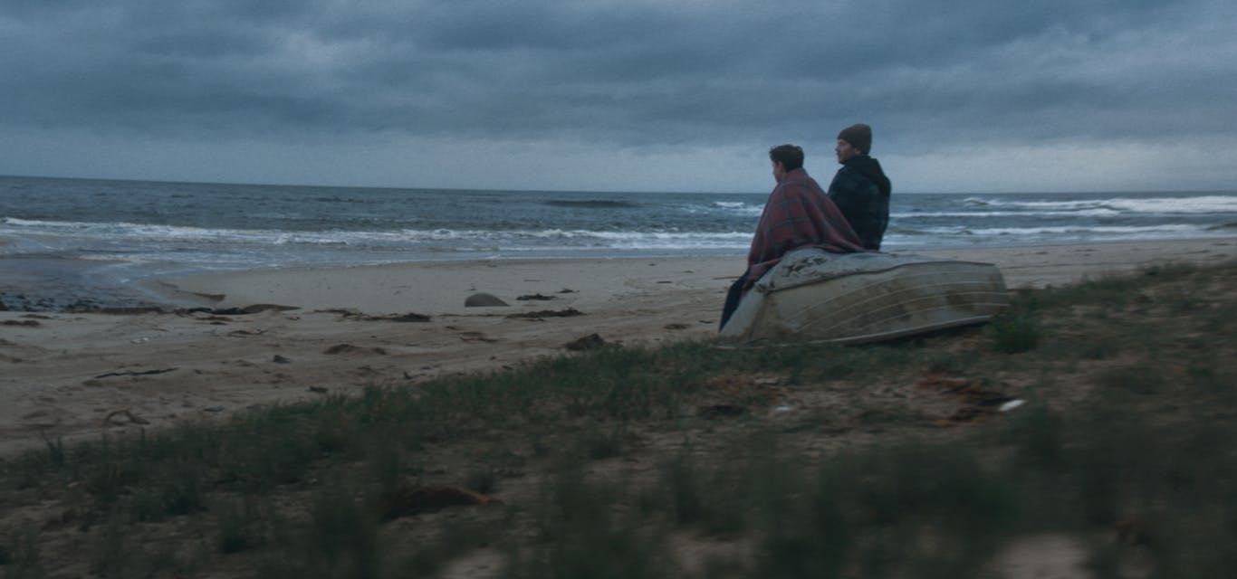 For Each Other A man and a boy sit on an upturned dinghy on a wild, overcast beach