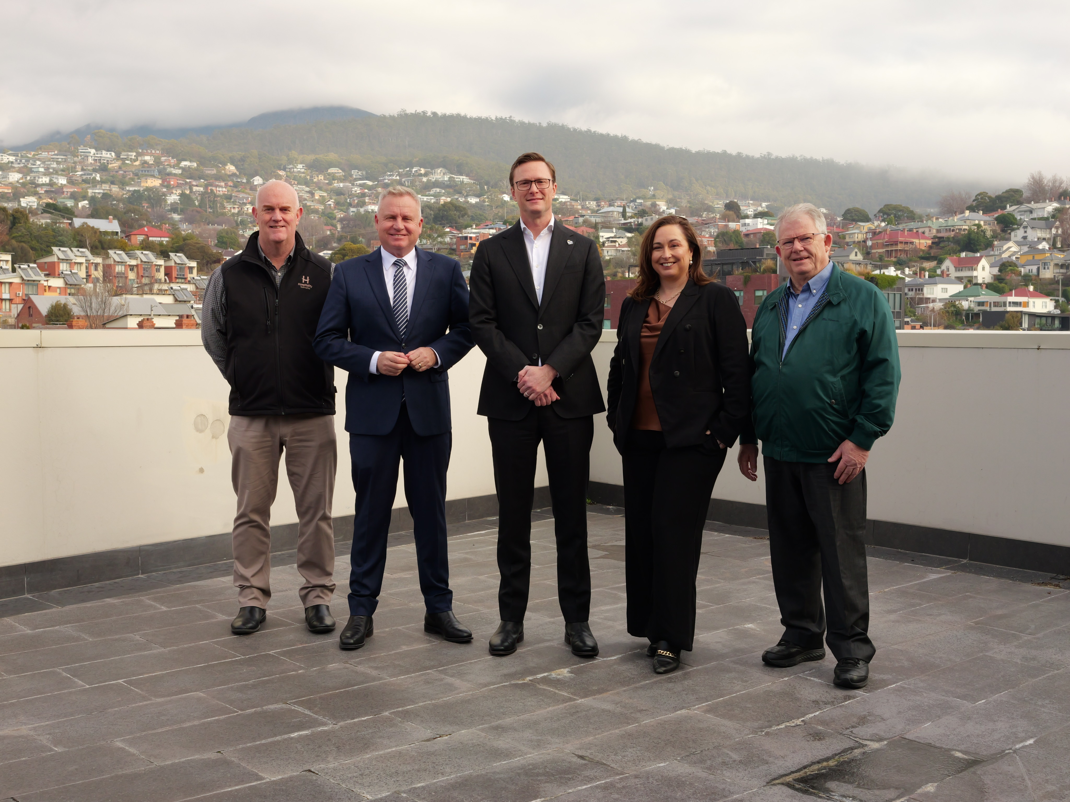 Four men and a woman stand on a corporate deck with suburbs and a mountain behind on an overcast day