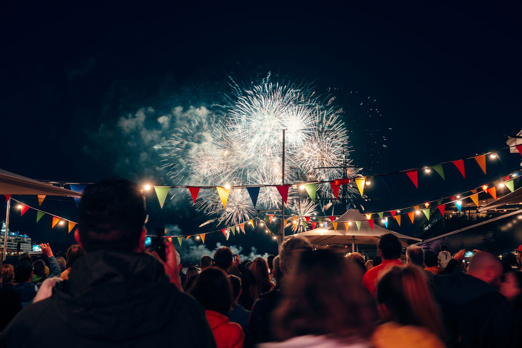 A crowd look at fireworks in the sky, with rainbow bunting