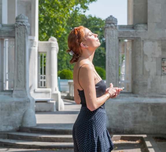 A white woman with red hair turns her face to the sun, eyes closed. Her hands are holding a cellphone in front of her, and there are stone columns in the background. 