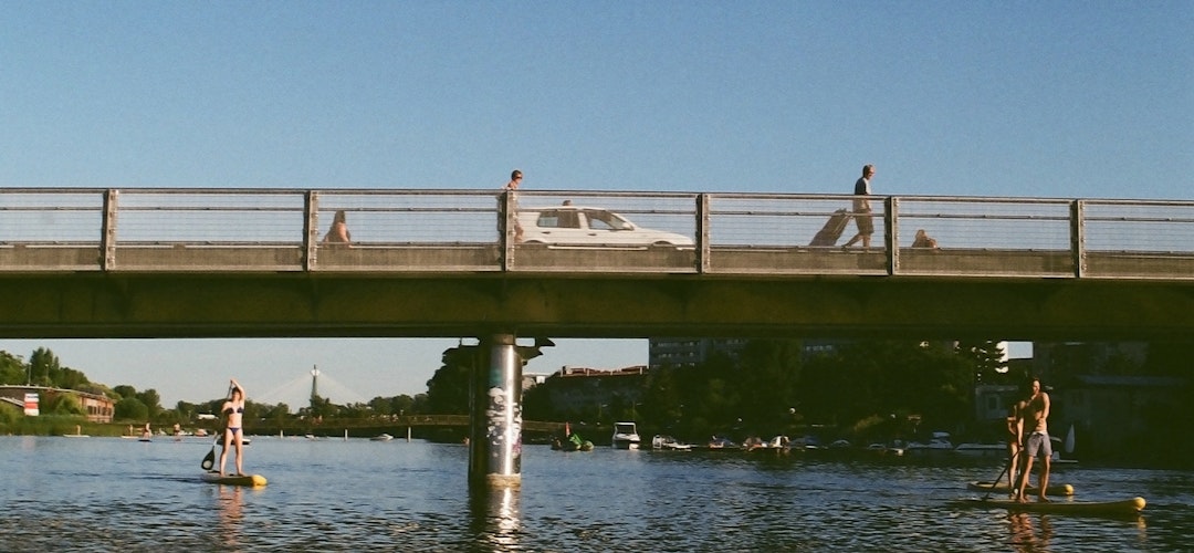 A low metal bridge covers a wide waterway. Two people in swimwear hold paddles as they use stand-up paddleboards beneath the bridge. 