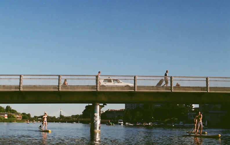 A low metal bridge covers a wide waterway. Two people in swimwear hold paddles as they use stand-up paddleboards beneath the bridge. 